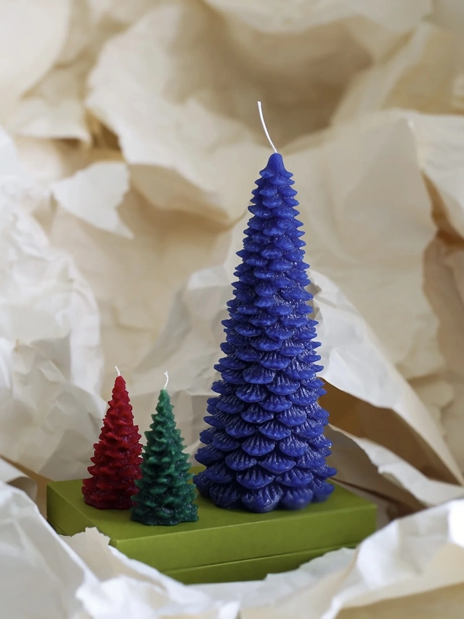 Large Christmas Tree Candle. a large indigo blue christmas tree shaped candle stands on a green box, nestled among crumpled cream tissue paper. a smaller red and a smaller dark green tree candle sit in front of it for scale, both with visible wicks. the textured, layered pine branch design and slightly variegated wax give all three candles a realistic, hand-poured finish.
