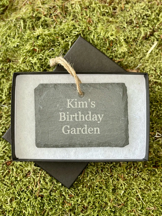 Engraved birthday message on a hanging slate sign in a gift box on a bed of moss.