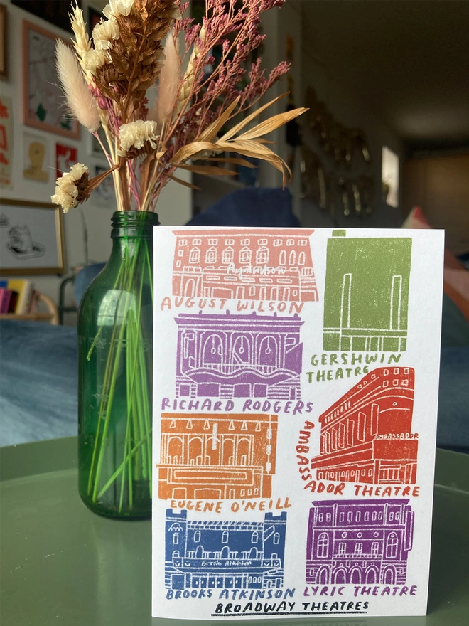 Greetings card with illustrated Broadway theatres, placed on a table with flowers in the background