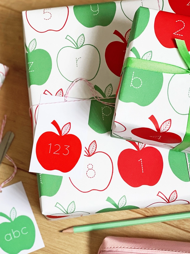 A gift wrapped in red and green apple wrapping paper, with letters and numbers inside each apple, is on a wooden surface.