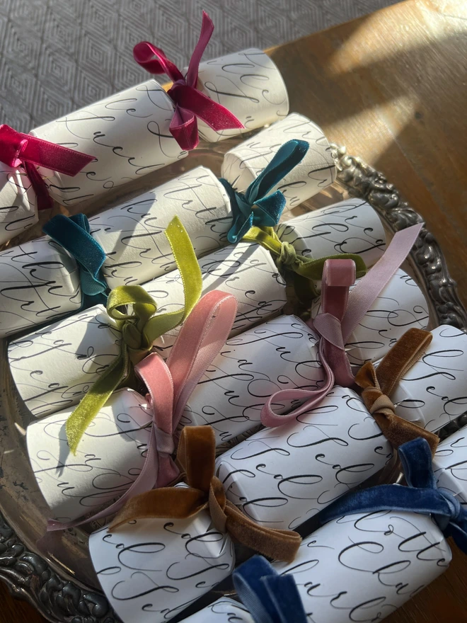 Selection of crackers personalised with calligraphy names tied with green, pink, mustard, blue, teal, rose, cerise velvet ribbon, laid on a silver tray on a wooden table