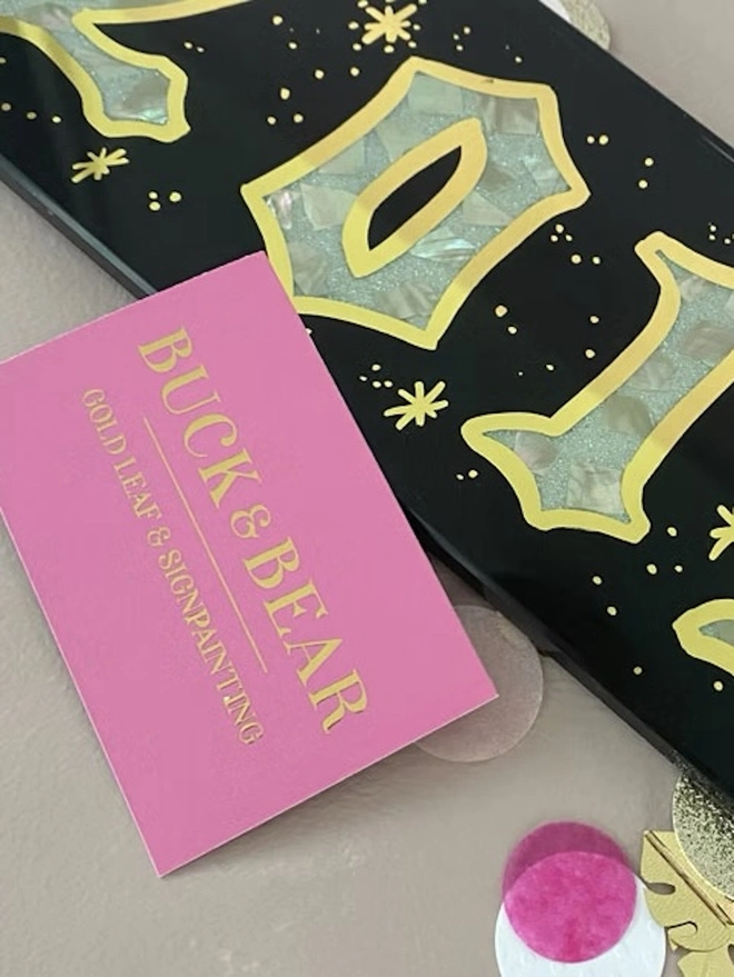 Personalised Year Date Sign, a black and gold personalised year sign sitting on a plain surface. It is joined by a small pink card. 