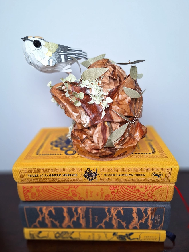 Handmade paper sculpture depicting a firecrest bird perched on a tree stump. The sculpture sits on a pile of books.
