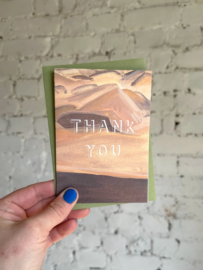 a greetings card featuring an illustration of a sand dunes landscape with the text “thank you” with a green envelope