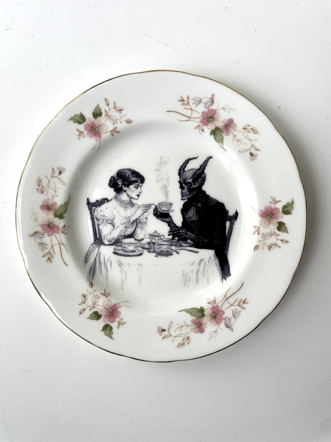 a vintage plate with an ornate edge, in the centre is a vintage illustration of a victorian lady drinking tea with the devil
