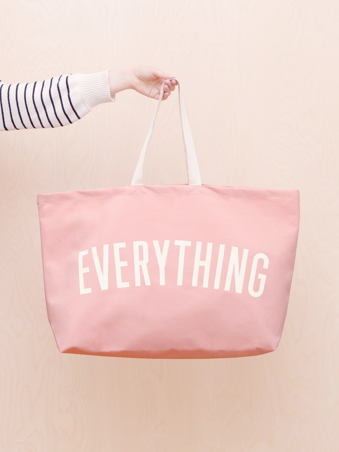 Everything oversized canvas tote bag in Pink held out in front of a plywood wall