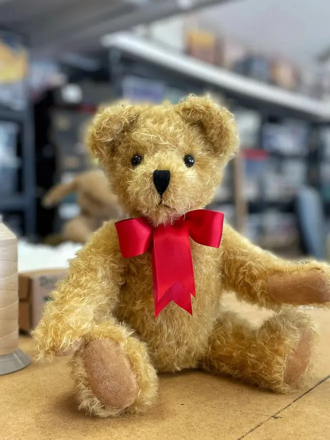 Handmade brown teddy bear sitting upright with a red ribbon bow against a kitchen background. 