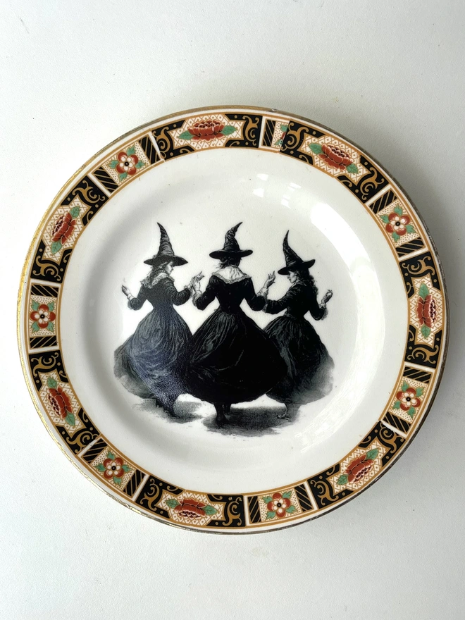 a coven of 3 witches dance hand in hand, the image is victorian in black and white and is printed directly onto an ornate vintage plate
