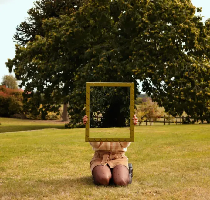 Girl holding a scallop wood mirror with a scalloped wooden frame in an open green area with a tree in the background.