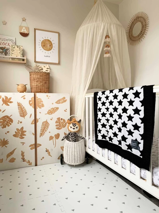 A neutral scandi style nursery with lots of natural wood, wicker accessories and a white cot. Over the cot is draped a black and white star blanket with a black trim.