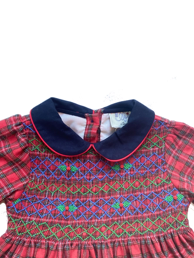 A red tartan dress with a navy collar and hand smocked detailing featuring holly embroidery