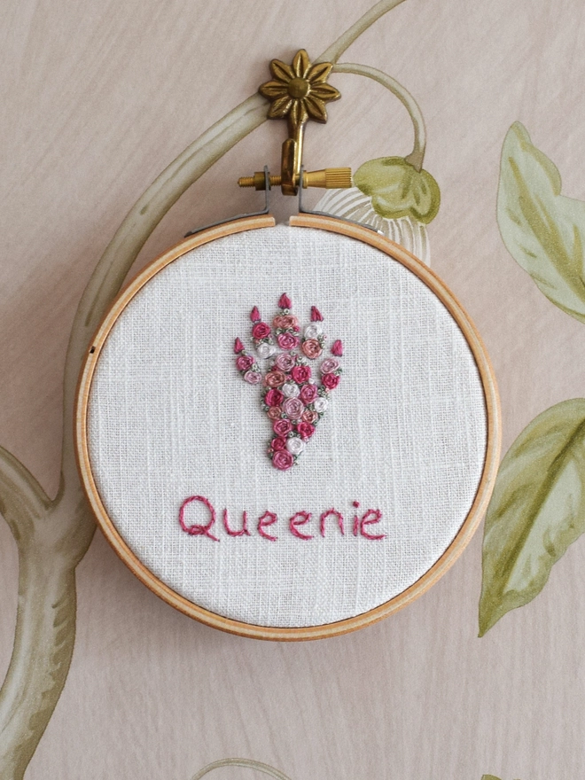 An embroidered Rabbits’ Foot, of woven wheel roses in 5 shades of pink with French Knot green grass.  Displayed in a hoop frame on a brass floral wall hook.