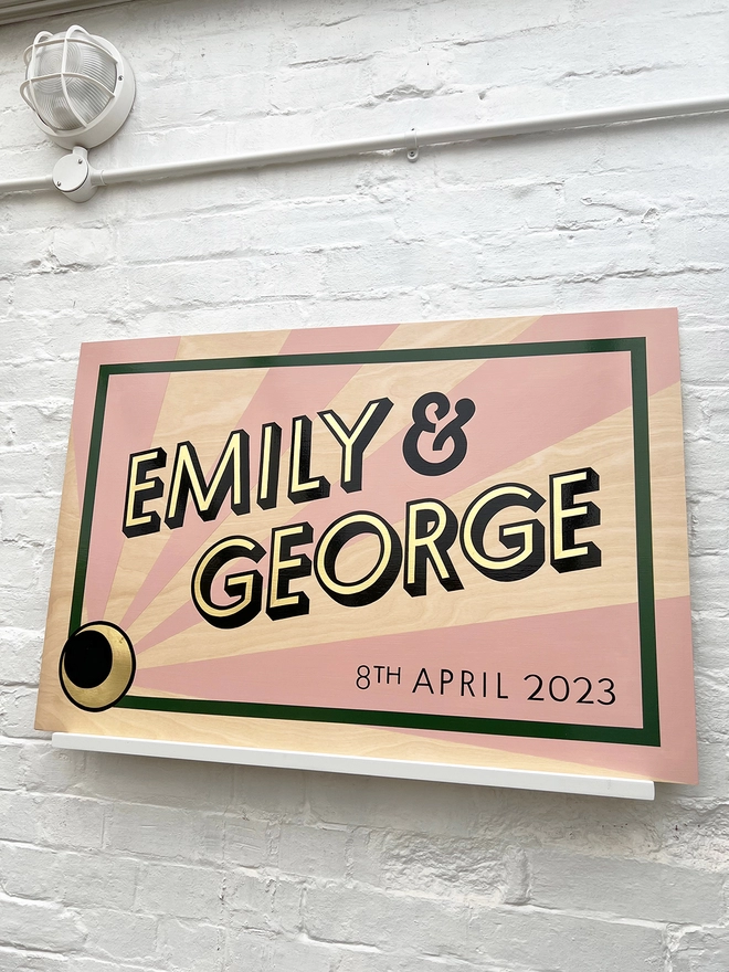 Modern wedding sign featuring the couple's names in gold leaf, outlined in black, and the wedding date, with a sunburst background and a border. Shown against a white brick wall, at an angle. 