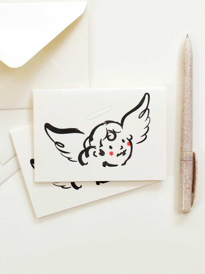 Christmas cards, envelopes and a pen pictured together in a group.  Cards feature a cute cherub character in black ink with red cheeks. 