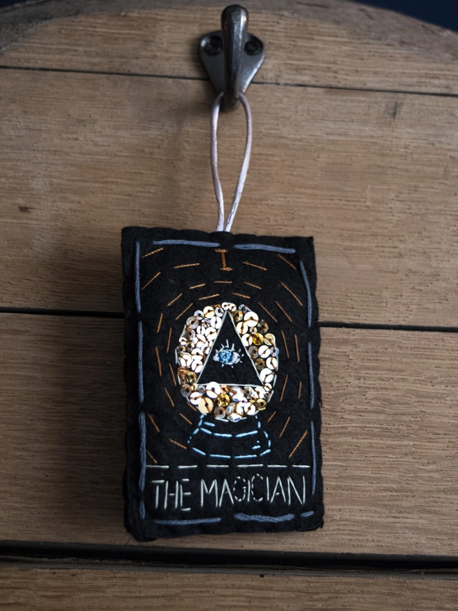 A handcrafted felt ornament with sequins and embroidery depicting "The Magician" tarot card. Colourful embroidery with an eye inside a crystal ball. 