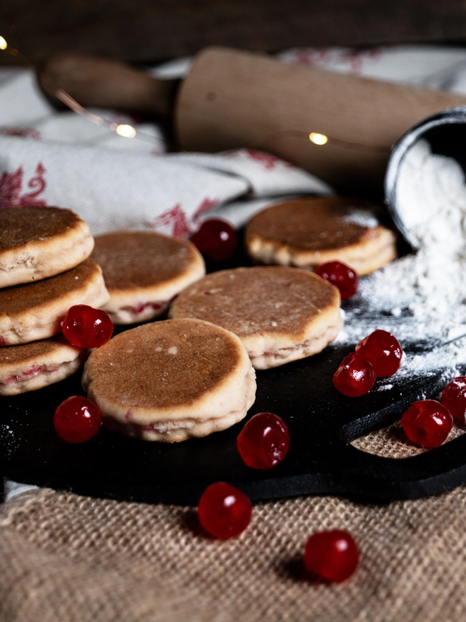 Cherry Bakewell Welsh Cakes on a black griddle bakestone with split flour, cherries and a rolling pin