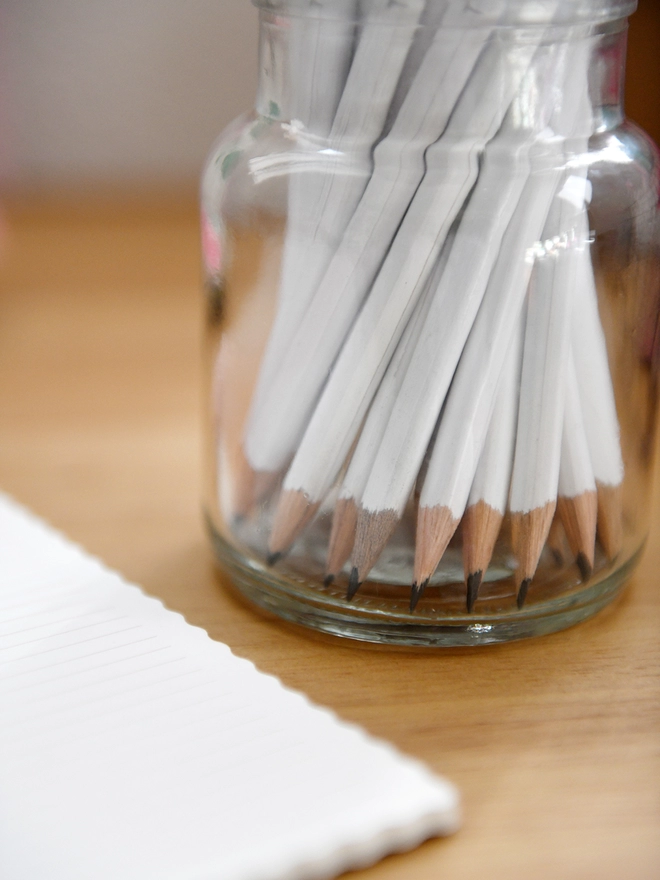 A glass jar full of white pencils with sharpened tips stands on a wooden desk.