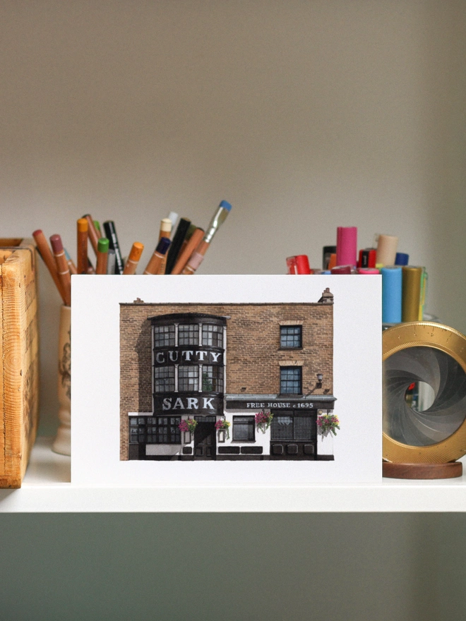 A watercolor print of the Cutty Sark pub displayed on a shelf among various art supplies