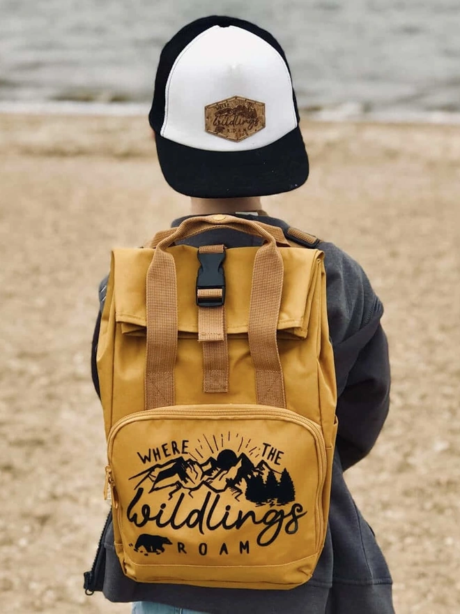 'Wildings' Children’s Recycled Roll Top Back Pack, a child sitting on the shore with a colourful backpack. 