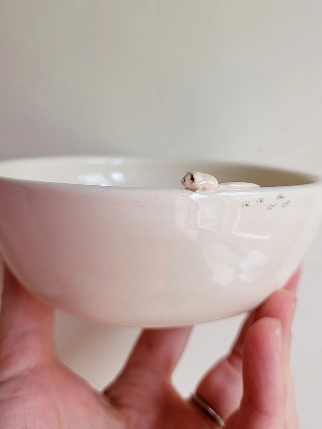 ivory white pottery cat bowl held in a bowl with a tiny mouse attached to the rim and tiny foot prints behind it on the bowl