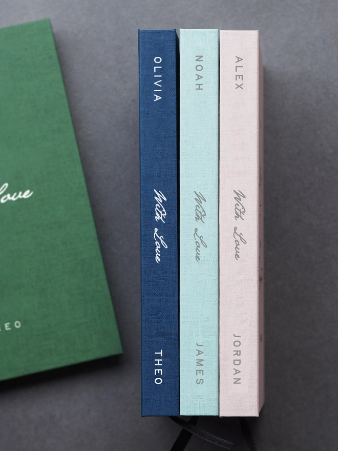 Personalised Hardcover With Love Notebook. The spines of three journals in different colours, personalised with a name at the top and at the bottom of the spine, with With Love written in the middle
