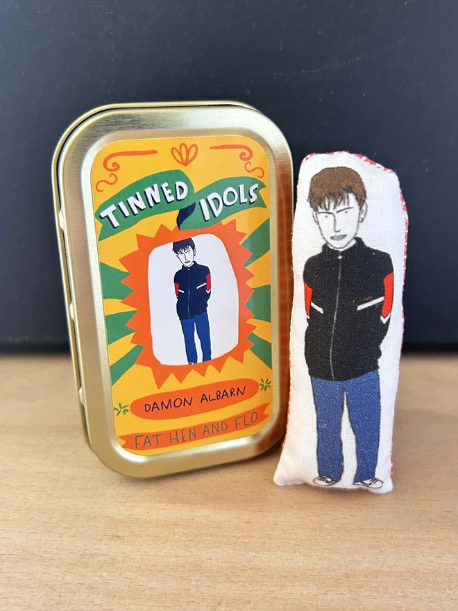 Mini fabric doll featuring illustration of Blur frontman, Damon Albarn, and gold tin.