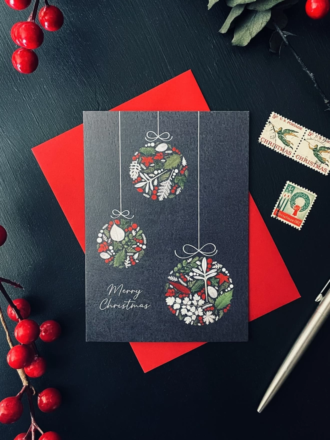 Christmas Card with Bauble Design Made from Pressed Winter Leaves, Holly, and Ivy on Red Envelope - Dark Charcoal Desk Setting