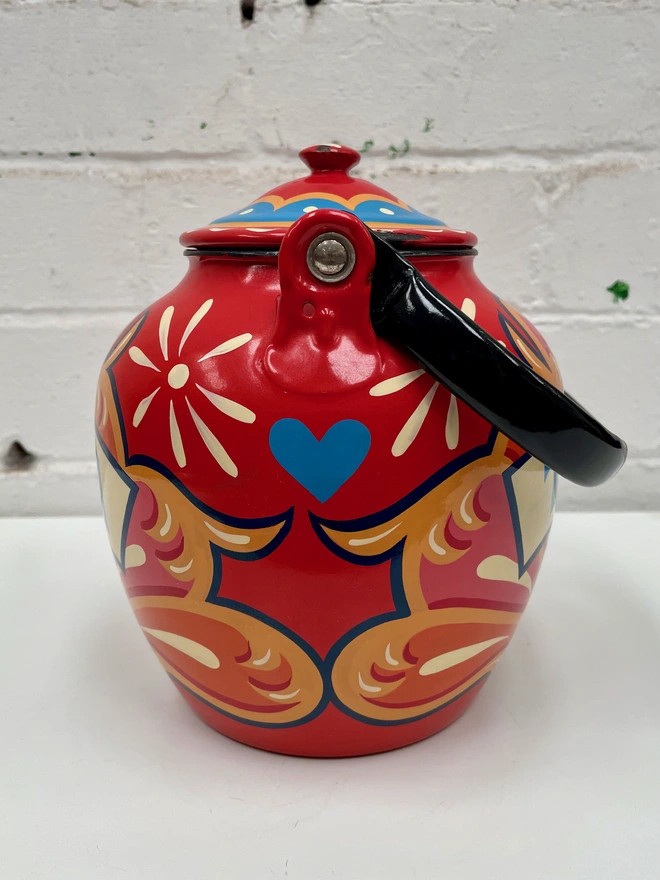 Red vintage enamel teapot with folding pouring handle, hand-painted with a traditional fairground inspired design in pink, orange and yellow. 'Tea' is painted in a banner across the centre.