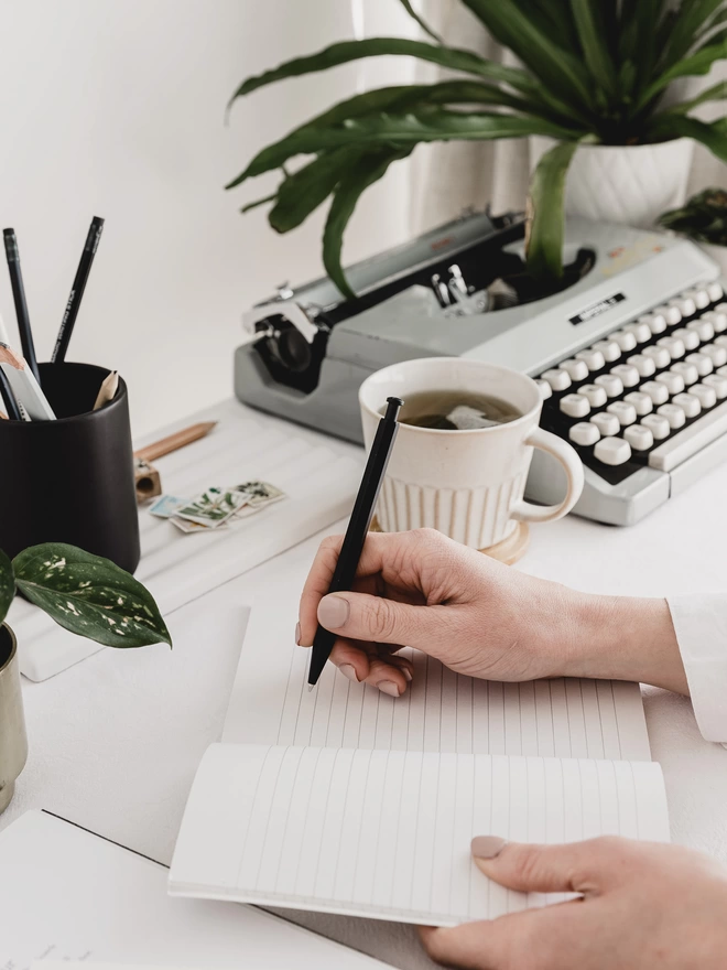 writing in a lined notebook on a desk with a typewriter