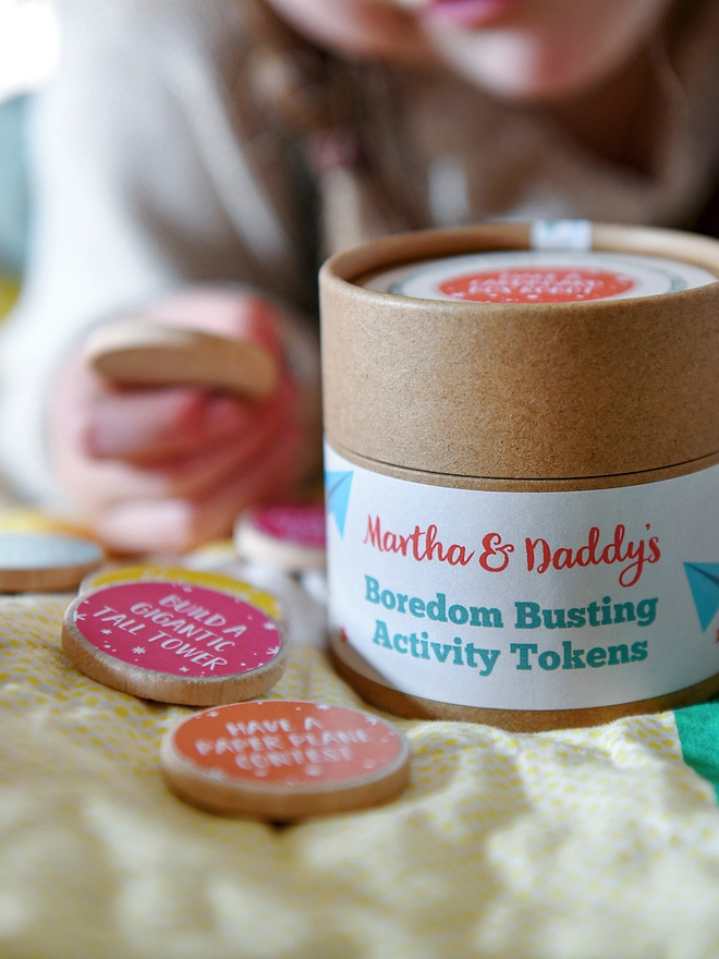 A cardboard jar with a label that reads Boredom Busting Activity Tokens, rests on a child's knee. Several wooden tokens with bright coloured labels lay beside it.
