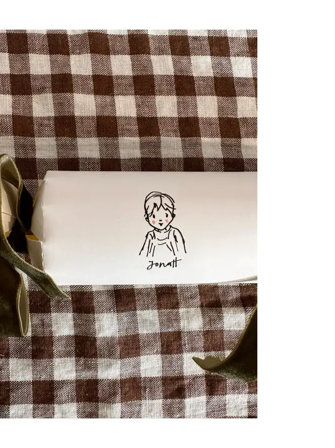 white christmas crackers, each with a bespoke illustration of a child or pet, with different coloured velvet ribbon. sitting on brown and white gingham table cloth