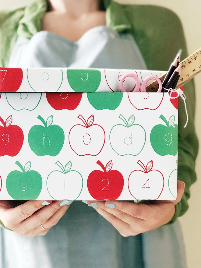 A gift wrapped in red and green apple wrapping paper, with letters and numbers inside each apple, is being held.