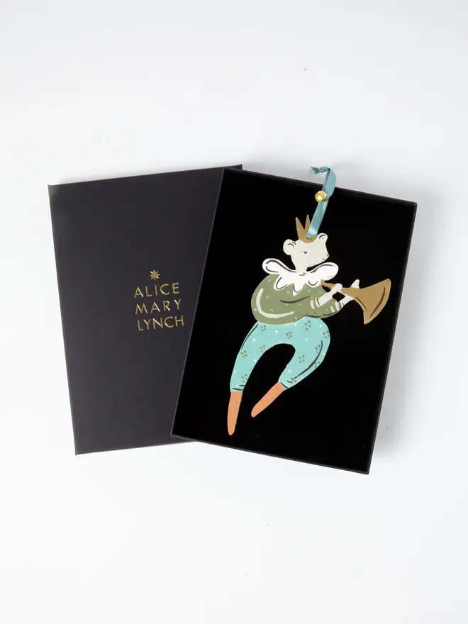 Mouse Herald Tree Dancer. the mouse herald tree dancer hanging ornament in olive green and teal tones sits inside its black presentation box, with the lid open to reveal "alice mary lynch" embossed in gold lettering. the mouse figure wears a gold crown, a ruffled white collar and a spotted olive jacket, playing a gold trumpet, with teal trousers ending in orange boots. the branded box gives the piece a polished, gift-ready presentation.