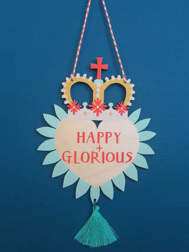 'Happy + Glorious' is hand printed on a colourful wooden heart with party crown and tassel. 