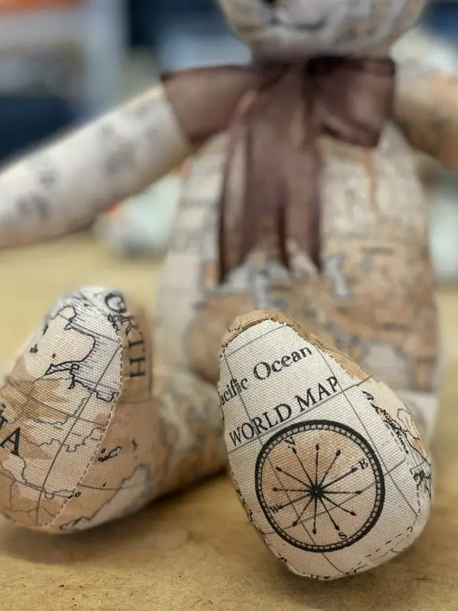 Teddy bear made from map-print fabric sitting upright on a wooden table, wearing a brown bow.