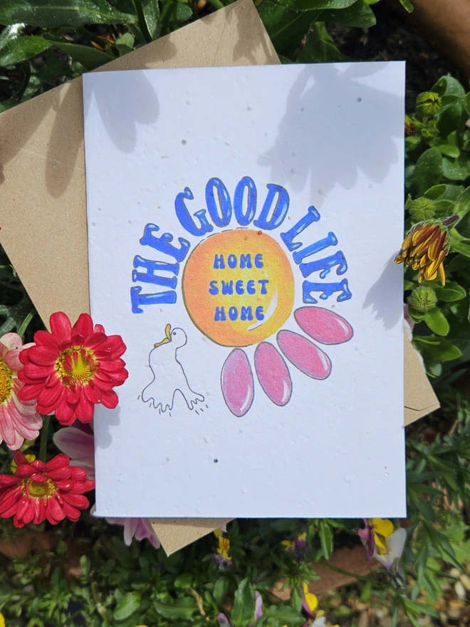 A white seed card with an illustration of a pink flower and a white bird. The Good Life is written using a retro font style in blue. In the centre of the flower it says Home Sweet Home. 