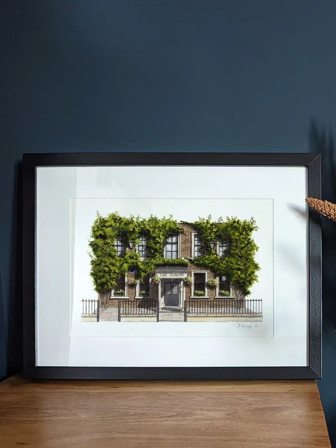A framed watercolor print of The Albion displayed on a wooden surface next to a vase of dried flowers.
