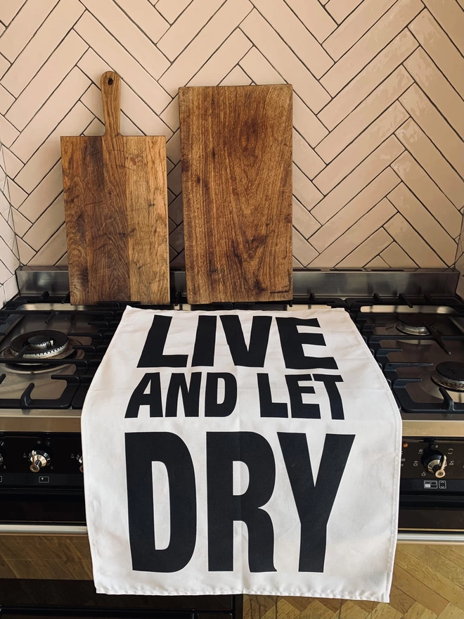 London Drying Live and Let Dry black screen printed text on white tea towel laying on hob/cooker with 2 wooden chopping boards leaning against tiled wall in background