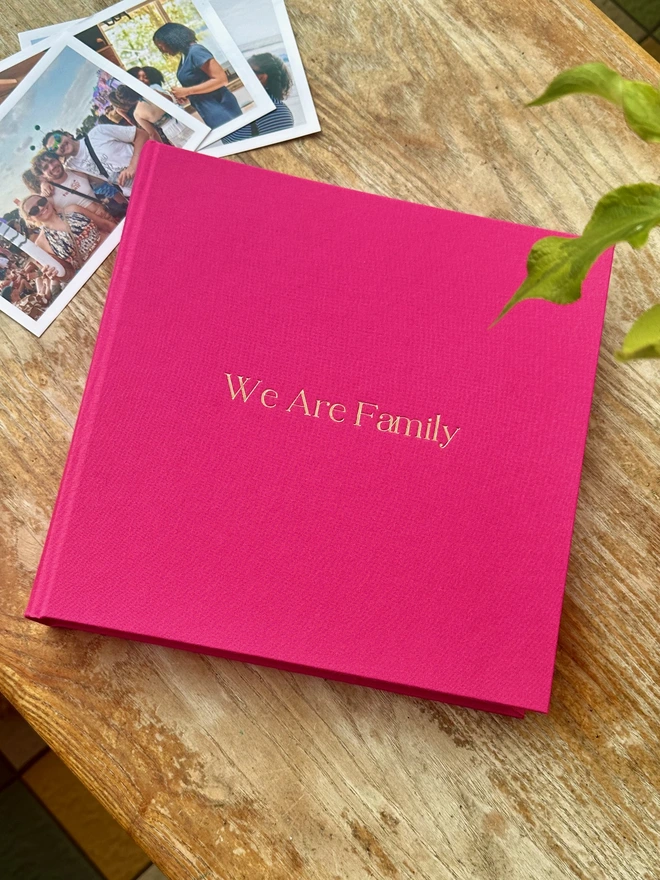 a pink photo album is on a wooden table, the album has been printed on the front the words We are Family. The album is surrounded by photos of family and friends