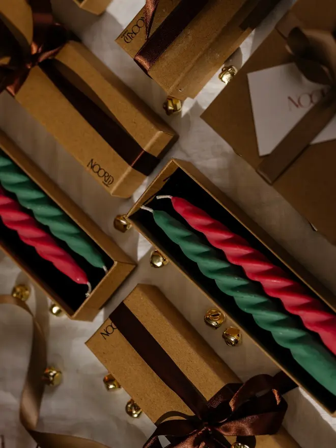 Christmas Spiral Candles Gift Box (Set Of 2). Pairs of red and green twisted taper candles in open kraft Noord gift boxes, surrounded by boxes tied with brown satin ribbon, gold jingle bells and a linen cloth. The gift-ready spiral candles make a festive present or a colourful finishing touch for Christmas tables.