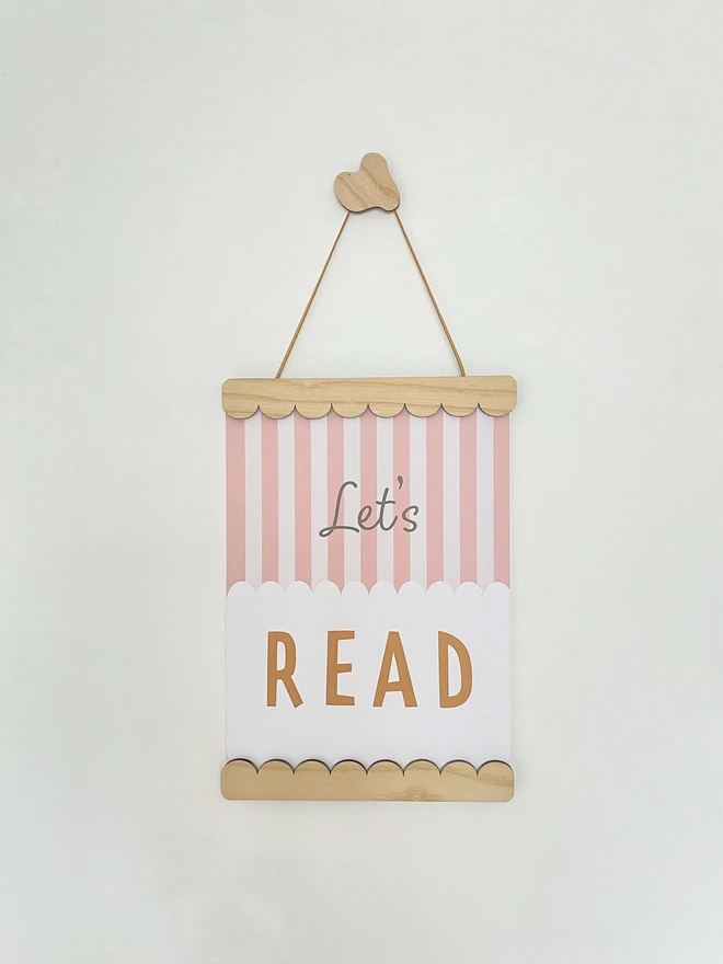 A wooden magnetic picture frame, strung with vegan suede. Hung on a wavy wooden wall dot. Displaying a colourful stripy kids print that says Let’s Read