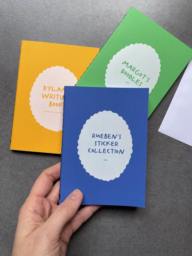 Three personalised brightly coloured pocket size notebooks on a dark worktop, one of the books says Reuben's sticker collection, one says Margot's Scribbles and the other says Dylan's writing book.