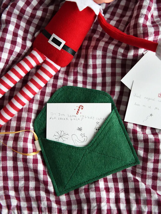 A handmade green Christmas envelope with a festive card tucked inside, written with a note to the elves, lays on a gingham cloth next to a Christmas elf toy.