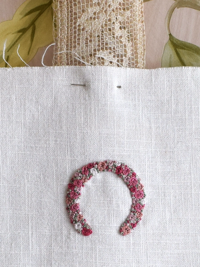 An embroidered Horseshoe, of woven wheel roses in 5 shades of pink with French Knot green grass.  Pinned to a length of vintage lace, with ends pointing down to allow Luck to flow over all who pass by.