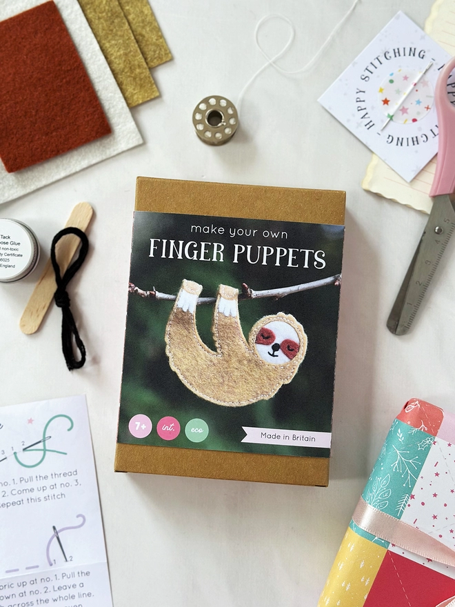 A small craft kit box to make a felt sloth finger puppet, lays on a white desk. Various craft kit contents lay around the box.