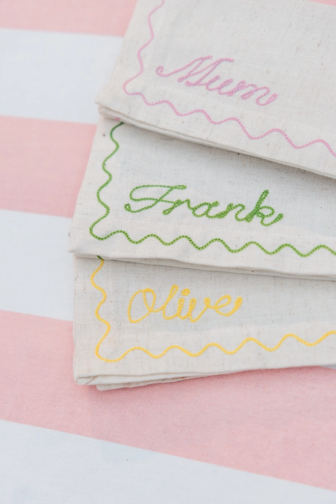 Personalised linen napkins with moss green, dusky pink and yellow embroidery - 'Mum, Frank & Olive' fanned out on a pink and white striped tablecloth