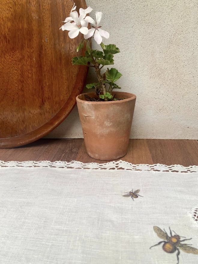 An upcycled vintage linen and lace table runner with printed bees and a potted white pelargonium