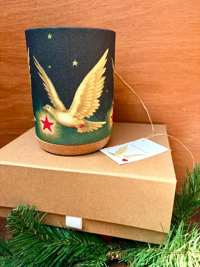 A festive candle shade with flying dove and stars, standing on a kraft card gift box