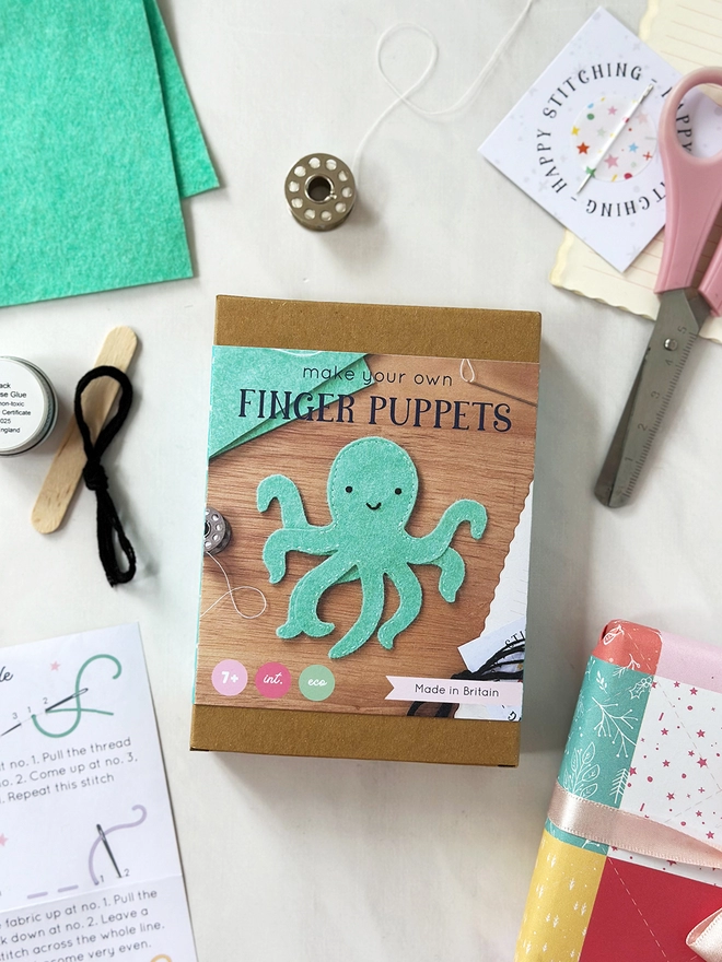 A small craft kit box to make a felt octopus finger puppet, lays on a white desk. Various craft kit contents lay around the box.