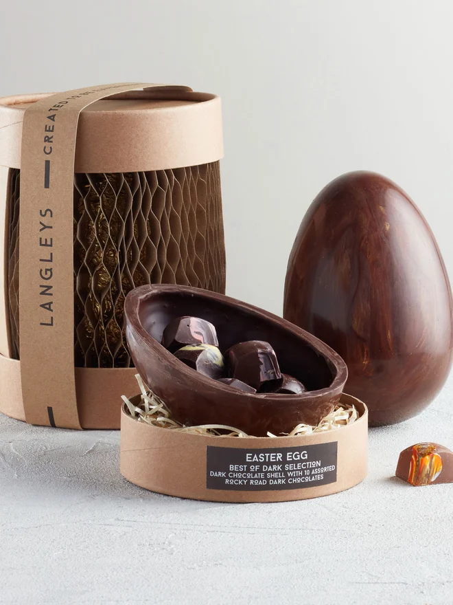 Best Of Dark Chocolate Easter Egg, a chocolate egg sitting on a white surface. It is accompanied by another chocolate egg that is opened showing the chocolates within. 